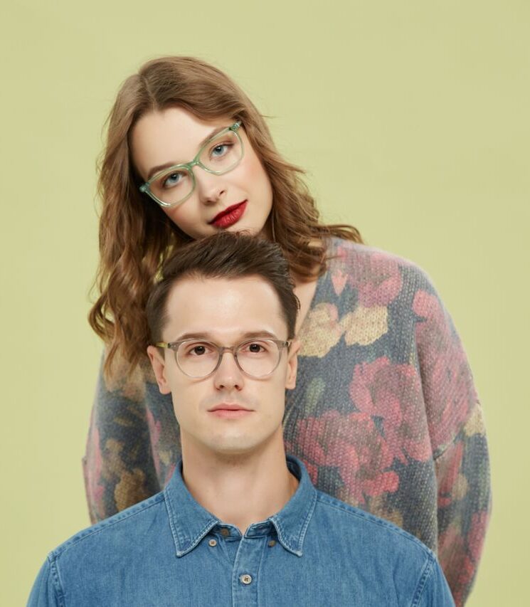A fashionable couple sporting trendy eyeglasses poses against a light background.